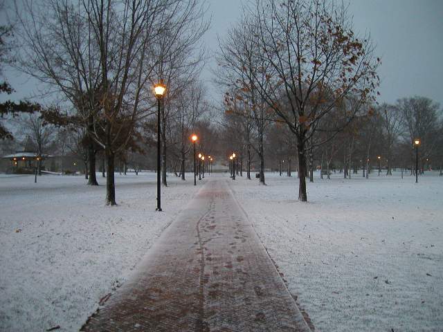 A brick path with a light dusting of snow and footprints, with grass and trees in a park on either side