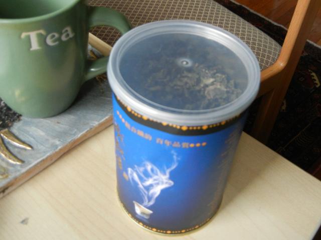 A blue canister with a clear plastic lid, containing loose tea, and a mug labelled tea, on a desk, with a chair