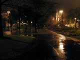 Dimly lit campus photo, with an asphalt walkway, wet from rain, some orange street lights, and a church tower visible in the background