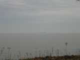 A distant barge on lake erie, gray water, party cloudy sky, and dead, weedy plants in the foreground