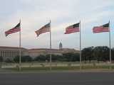 Four American flags flying on flag poles with a large government-looking building in the distant background, and some trees