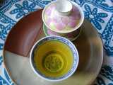 A gaiwan, a Chinese lidded bowl, closed, with a pink and yellow pattern on the lid, and a small teacup with a rich golden-yellow infusion in it