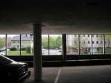 Photo taken from inside a parking garage, mostly dark, car on the left, looking out to a restrained view of a commercial building on the right and a house on the left