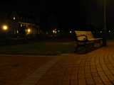 A brick sidewalk, wooden bench with metal arms, and some pretty brick buildings in the background, on a college campus, at night