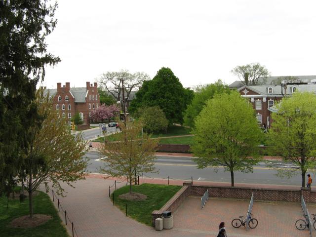 A street intersection with numerous trees leafing out in early spring, in the middle of a college campus, with brick buildings, sidewalks, and a brick terrace