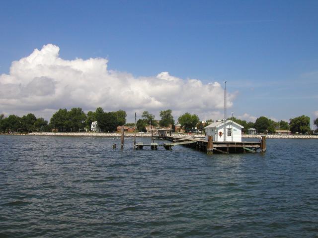 A pier in water with a small white building on it, coming out from land with rocks along the edge, some trees, and a brick building