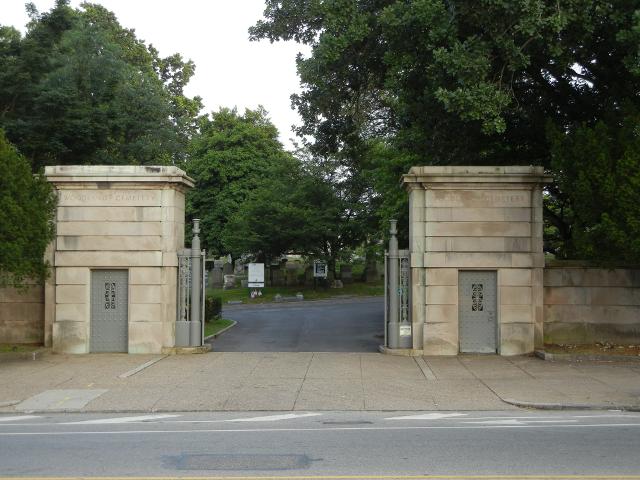 Two large stone columns with metal doors in them, with an open gate leading into a cemetery with lush, green trees, a sidewalk and street in front
